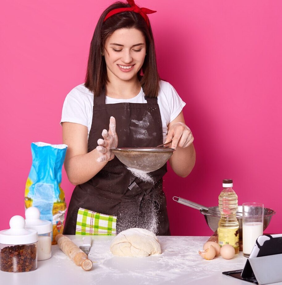 portrait-skillful-talented-cook-putting-flour-through-sieve-half-ready-pie-with-raisin-brunette-cute-young-model-poses-isolated-bright-pink-baking-cooking-concept_176532-8425